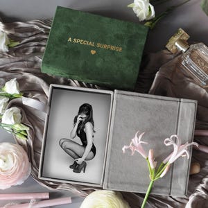 K&ouml;nnte beinhalten: Eine gr&uuml;ne Samtbox mit dem goldenen Text "A SPECIAL SURPRISE" und einem Herz. Die Box ist ge&ouml;ffnet und zeigt ein Schwarz-Wei&szlig;-Foto einer Person. Das Foto befindet sich in einer grauen Box. Blumen und Parf&uuml;m sind ebenfalls im Bild.