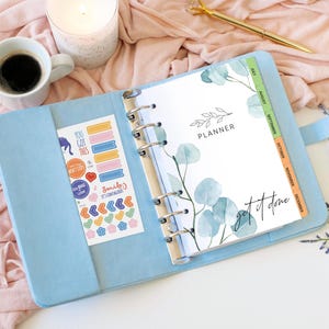 May include: A light blue six-ring binder planner featuring a white cover page with green eucalyptus leaf illustrations, the word PLANNER, and the text get it done. A sticker sheet with colorful icons and phrases is tucked into the left pocket.