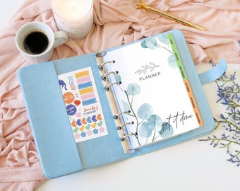 May include: A light blue six-ring binder planner featuring a white cover page with green eucalyptus leaf illustrations, the word PLANNER, and the text get it done. A sticker sheet with colorful icons and phrases is tucked into the left pocket.