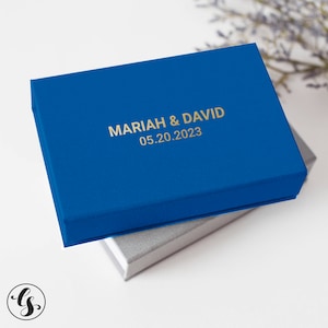 May include: A cobalt blue box with gold lettering that reads "MARIAH & DAVID" and the date "05.20.2023". The box is sitting on top of a silver box. The background is white with a hint of purple flowers.