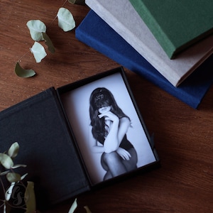 K&ouml;nnte beinhalten: Eine schwarze Fotobox mit einem Schwarz-Wei&szlig;-Foto einer Person mit Spitzenmaske. Drei Hardcover-B&uuml;cher in Blau, Grau und Gr&uuml;n sind im Hintergrund gestapelt. Eukalyptusbl&auml;tter sind verstreut.