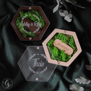 Puede incluir: Dos cajas de anillos hexagonales con tapas. La caja superior est&aacute; hecha de madera oscura con una tapa transparente y presenta un lecho de musgo verde con un soporte de anillo de madera grabado con "Ashley & Riley". La caja inferior est&aacute; hecha de madera clara con una tapa transparente y presenta un lecho de musgo verde con un soporte de anillo de madera grabado con "04.10.2023". La tercera caja est&aacute; hecha de acr&iacute;lico transparente con forma hexagonal y presenta un dise&ntilde;o floral con la palabra "Theis" grabada en el centro.