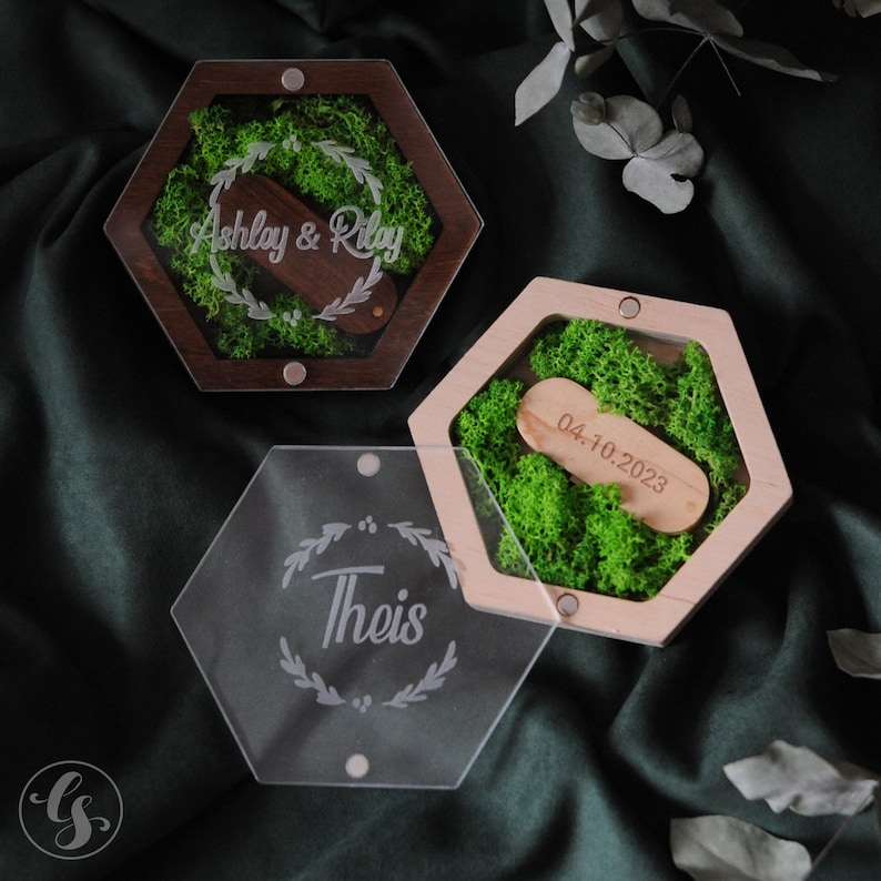 Puede incluir: Dos cajas de anillos hexagonales con tapas. La caja superior est&aacute; hecha de madera oscura con una tapa transparente y presenta un lecho de musgo verde con un soporte de anillo de madera grabado con "Ashley & Riley". La caja inferior est&aacute; hecha de madera clara con una tapa transparente y presenta un lecho de musgo verde con un soporte de anillo de madera grabado con "04.10.2023". La tercera caja est&aacute; hecha de acr&iacute;lico transparente con forma hexagonal y presenta un dise&ntilde;o floral con la palabra "Theis" grabada en el centro.