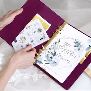 May include: A deep purple fabric wedding planner with gold ring binding. The open planner displays a white page with green eucalyptus illustrations, gold geometric lines, and the text "OUR together IS forever WEDDING PLANNER". A sticker sheet is tucked inside.