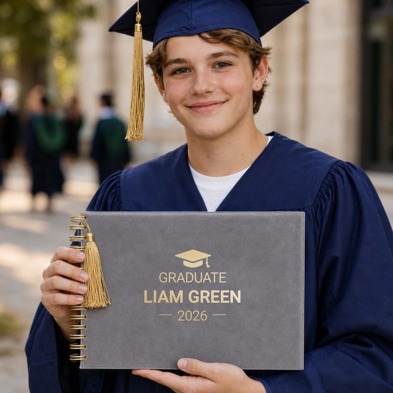May include: A graduate in a navy blue cap and gown holds a gray diploma case with gold text that reads "GRADUATE LIAM GREEN - 2026". The diploma case has a gold tassel and spiral binding.