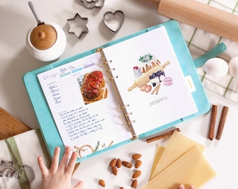 Puede incluir: Un archivador de recetas azul claro con anillos dorados, que muestra una receta de tarta de manzana y una ilustración de postres. Utensilios para hornear, como un rodillo, cortadores de galletas y palitos de canela, están dispuestos alrededor.