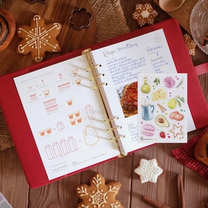 May include: A red ring-bound recipe planner open to a page with kitchen measurement conversions and handwritten notes, featuring a sticker sheet with watercolor food illustrations including a mug, avocado, and macaron, surrounded by gingerbread cookies.