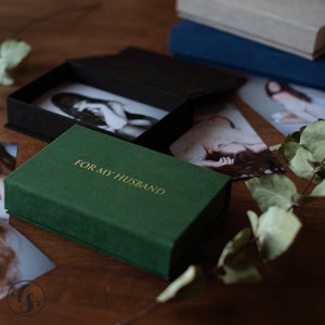 K&ouml;nnte beinhalten: Eine dunkelgr&uuml;ne Schachtel mit dem goldenen Text "FOR MY HUSBAND" befindet sich im Vordergrund. Die Schachtel ist ge&ouml;ffnet und zeigt eine schwarze Schachtel mit einem Schwarz-Wei&szlig;-Foto. Weitere Fotos und B&uuml;cher befinden sich im Hintergrund.