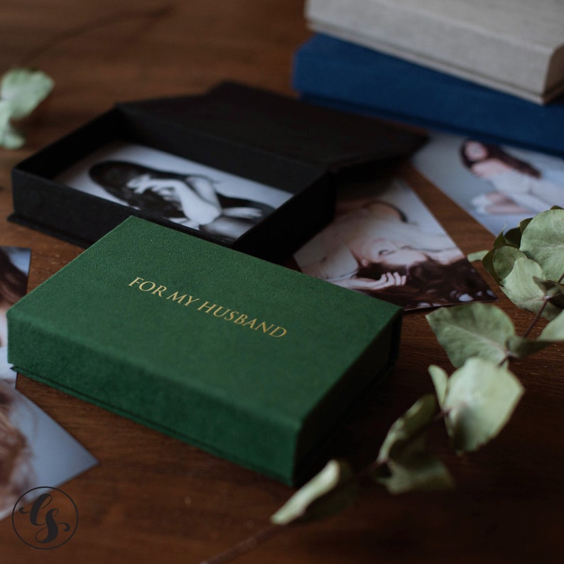 K&ouml;nnte beinhalten: Eine dunkelgr&uuml;ne Schachtel mit dem goldenen Text "FOR MY HUSBAND" befindet sich im Vordergrund. Die Schachtel ist ge&ouml;ffnet und zeigt eine schwarze Schachtel mit einem Schwarz-Wei&szlig;-Foto. Weitere Fotos und B&uuml;cher befinden sich im Hintergrund.