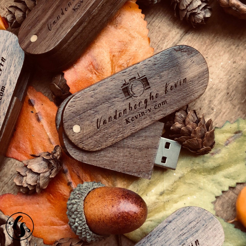 Puede incluir: Una memoria USB de madera con forma de coraz&oacute;n con un icono de c&aacute;mara y el texto "Vandenberghe Kevin kevin-v.com" grabado en la parte delantera. La memoria USB est&aacute; sobre un lecho de hojas de oto&ntilde;o.