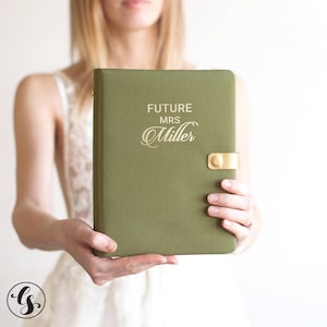 May include: An olive green fabric-covered planner featuring the gold-colored text "FUTURE MRS Miller" printed on the front cover. The planner includes a gold-colored metal snap closure on the right side to keep the cover securely fastened.