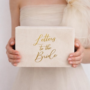 May include: An off-white rectangular box with the gold script text "Letters to the Bride." The box is held in front of a person wearing a cream-coloured dress. The box has a soft, textured appearance.