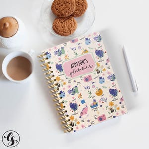 May include: A spiral-bound planner with a cream background, adorned with colorful cat illustrations and floral accents. The planner features the text "Addyson's planner" in a pink frame. A cup of coffee, cookies, and a pen are also visible.