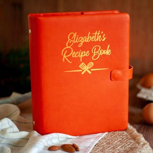 May include: A bright orange recipe book binder with a gold embossed inscription that reads Elizabeth's Recipe Book above a graphic of a spatula and whisk. The binder features a side strap closure and gold metal hardware on the spine.