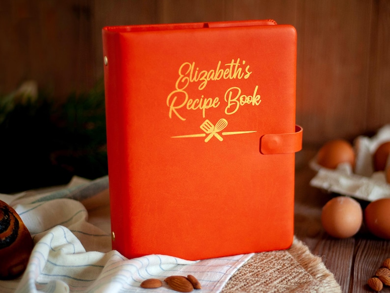May include: A bright orange recipe book binder with a gold embossed inscription that reads Elizabeth's Recipe Book above a graphic of a spatula and whisk. The binder features a side strap closure and gold metal hardware on the spine.