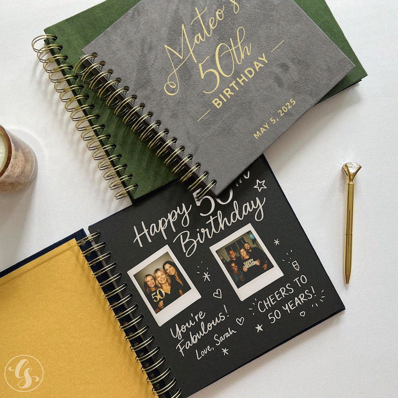 May include: A collection of spiral-bound notebooks with gold accents. One notebook is gray with "Mateo's 50th Birthday" in gold lettering. Another is black with "Happy 50th Birthday" and photos. A gold pen with a diamond-shaped top is also present.