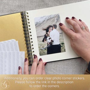 May include: An open scrapbook with a gold-colored spine and pages. A photograph of a couple is placed on a page. The photo is held in place by a person's hand with dark red nail polish. Additional photo corners are available.