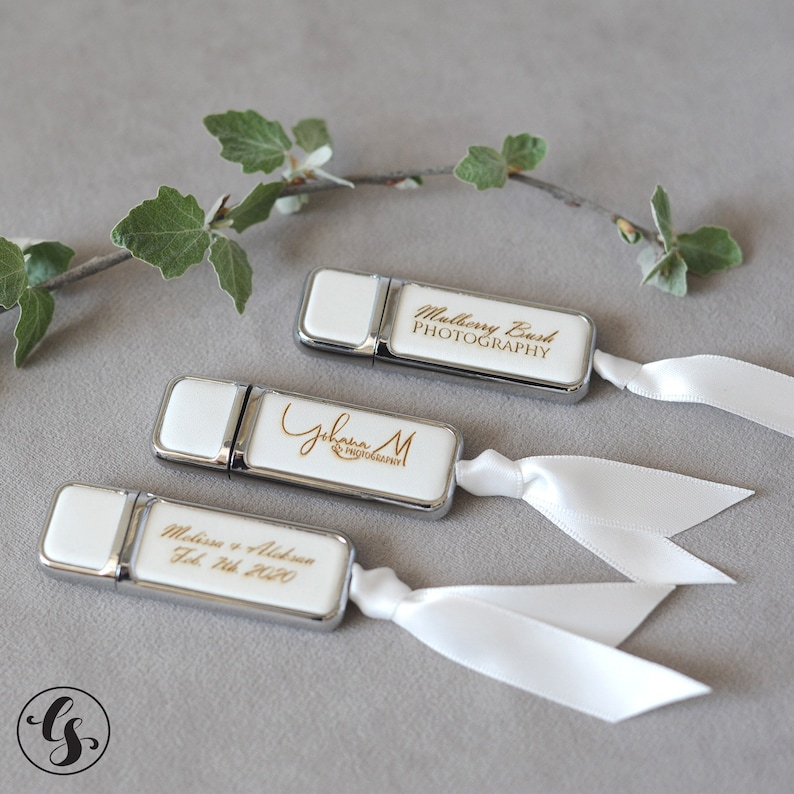 K&ouml;nnte beinhalten: Drei wei&szlig;e USB-Sticks mit silbernen Akzenten und wei&szlig;en B&auml;ndern. Die Sticks sind mit dem Text "Mulberry Bush PHOTOGRAPHY", "Johanna M PHOTOGRAPHY" und "Melissa & Aldren Feb 7th 2020" graviert.