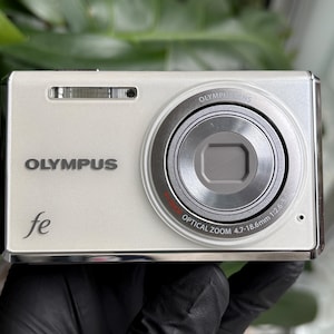May include: Off-white Olympus fe digital camera with a silver lens and trim. The camera has the word "OLYMPUS" printed on the front, along with the model name "fe". The lens has "OPTICAL ZOOM 4.7-18.6mm 1:2.6-5.0" printed on it.