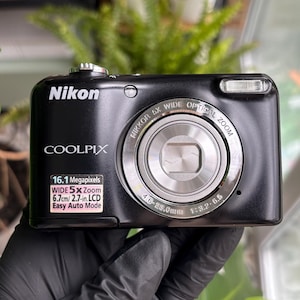 May include: Black Nikon Coolpix digital camera with a silver lens and flash. The camera has the text "COOLPIX" and "Nikon" printed on the front. It also has a sticker that says "16.1 Megapixels, WIDE 5x Zoom, 6.7cm/2.7-in. LCD, Easy Auto Mode".