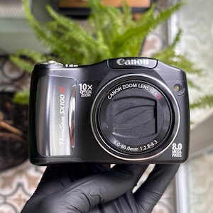 May include: A black Canon PowerShot SX100 IS digital camera featuring a 10x optical zoom lens with 6.0-60.0mm 1:2.8-4.3 specifications and 8.0 megapixels. The front of the camera displays the brand name, model, and zoom capabilities.