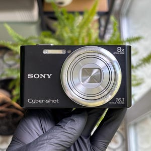 May include: A black Sony Cyber-shot digital camera featuring an 8x optical zoom lens, a built-in flash, and 16.1 megapixels printed on the front. The compact device has a silver lens ring and white text branding on the front face.