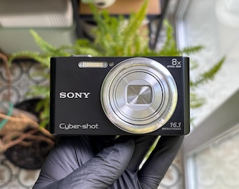 May include: A black Sony Cyber-shot digital camera featuring an 8x optical zoom lens, a built-in flash, and 16.1 megapixels printed on the front. The compact device has a silver lens ring and white text branding on the front face.