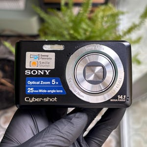 May include: A black Sony Cyber-shot digital camera featuring a 5x optical zoom, a 25mm wide-angle lens, and 14.1 megapixels. The front includes labels for Sweep Panorama, Smile Shutter, and the Sony Lens branding around the silver lens ring.