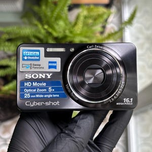 May include: A black Sony Cyber-shot digital camera featuring a Carl Zeiss Vario-Tessar lens, 5x optical zoom, 16.1 megapixels, and HD movie capabilities. The front panel displays labels for Optical SteadyShot, Sweep Panorama, and a 25 mm wide-angle lens.