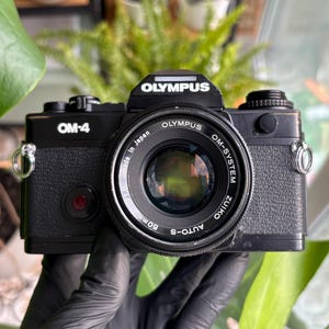 May include: Black Olympus OM-4 camera with a 50mm lens. The camera has the text "OM-4" and "OLYMPUS" on the body. The lens is labeled "OM-SYSTEM ZUIKO AUTO-S 50mm". The camera is a vintage film camera.