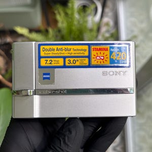 May include: A silver Sony Cyber-shot digital camera featuring a 3.0-inch hybrid LCD monitor, 7.2 megapixels, and Zeiss lens branding. The front panel displays text including Double Anti-blur Technology, Super SteadyShot, and Stamina battery life.
