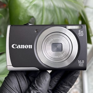 May include: A black Canon digital camera with a silver lens and trim. The camera has a 5x optical zoom and 16.0 megapixels. The Canon logo is visible on the front.