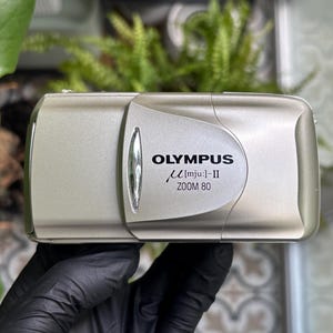 May include: A silver Olympus mju:-II Zoom 80 compact film camera with a sliding lens cover. The front of the camera features the brand name Olympus, the model name mju:-II, and the text Zoom 80 printed in black on the metallic silver body.