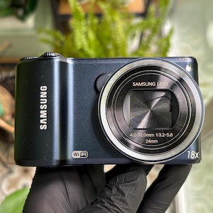 May include: A dark blue Samsung digital camera featuring a lens with 4.0-72.0mm 1:3.2-5.8 24mm specifications, an 18x zoom capability, and a Wi-Fi logo printed on the front body. The camera has a metallic finish and a retractable lens mechanism.