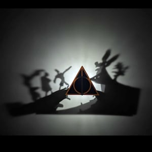 May include: A triangular wooden object featuring a circular and vertical line cutout design. The object casts a shadow on the wall that depicts silhouettes of three figures standing on a landscape with a tree and a large hand-like shape.