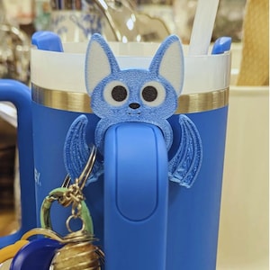 May include: A blue mug with a bat-shaped accessory on the handle. The bat has large eyes and wings. A keychain with various charms hangs from the handle. The mug has a white interior and a gold rim.