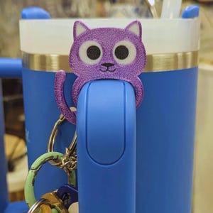 May include: A purple glitter cat charm is attached to a blue tumbler handle. The cat charm has large black eyes and a small nose. A keychain with various colored rings hangs below the charm.
