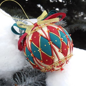 Peut inclure: Une boule de Noël décorative avec un motif rouge, turquoise et crème. L'ornement est orné de garnitures dorées, de rubans rouges et turquoise et de petits accents en forme d'étoile. Il est suspendu à une ficelle dorée.