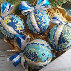 Op de afbeelding: Vier blauwe en witte kerstballen met gouden accenten en zilveren en blauwe strikken. De ballen zijn gemaakt van stof en hebben een gestikt ontwerp.