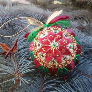 Op de afbeelding: Een handgemaakte kerstbal met een rood en groen ontwerp, versierd met gouden details en kralen. De bal heeft een sterrenpatroon en wordt opgehangen met een gouden draad en lint. Het ligt op blauwgroene dennennaalden.