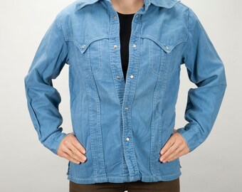 May include: A light blue denim shirt with a button-down front and long sleeves. The shirt has a classic Western style with two flap pockets on the chest.