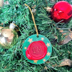 May include: A circular, green Christmas tree decoration featuring gold stars and a red wax seal design. The seal displays the letters "TM" in white. The ornament is suspended by a gold string, set against a green Christmas tree.