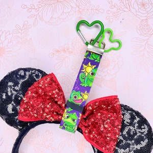 May include: Black sequined mouse ears with a red sequined bow. A purple lanyard with a cartoon design of a chameleon and a sun is attached to a green heart-shaped carabiner.