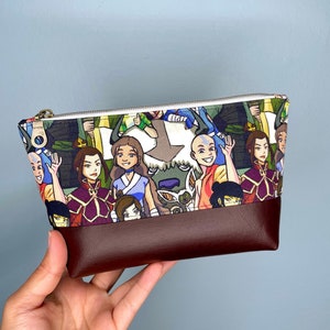 Puede incluir: Un neceser rectangular con cierre de cremallera. La parte superior presenta un estampado colorido de personajes animados, mientras que la parte inferior es de color marrón oscuro liso. La bolsa se sostiene en una mano.