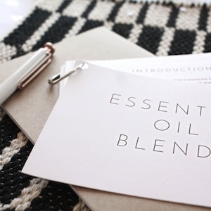 Könnte beinhalten: Eine weiße Seite mit dem Text "ESSENTIAL OIL BLENDS" in schwarzer Schrift. Die Seite liegt auf einem braunen Notizbuch mit einem weißen Stift und einer Sicherheitsnadel.