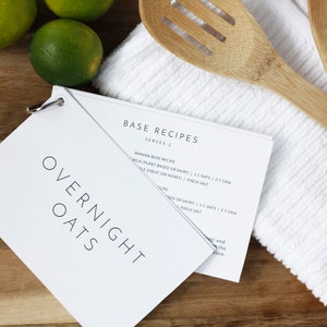 May include: A white booklet with the title "OVERNIGHT OATS" and a smaller booklet with the title "BASE RECIPES" on a wooden surface. The smaller booklet has a recipe for banana base overnight oats. There are two wooden spoons and a white kitchen towel on the surface.