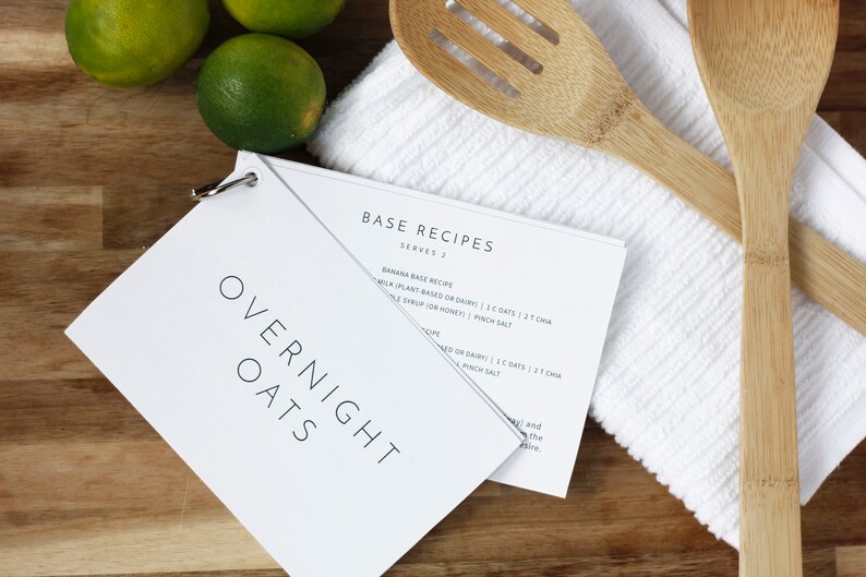 May include: A white booklet with the title "OVERNIGHT OATS" and a smaller booklet with the title "BASE RECIPES" on a wooden surface. The smaller booklet has a recipe for banana base overnight oats. There are two wooden spoons and a white kitchen towel on the surface.