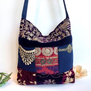 May include: A patchwork shoulder bag with a navy blue strap. The bag features a mix of fabrics in deep purple, red, and navy blue, embellished with gold embroidery and sequins. The bag has a bohemian style.