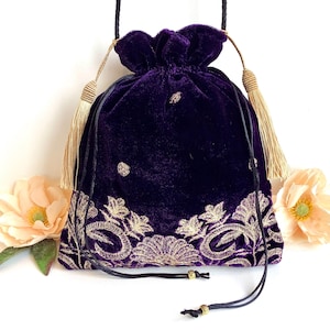 May include: A deep purple velvet drawstring bag with gold embroidered floral designs. The bag has a black drawstring closure and is adorned with gold tassels. The bag is decorated with small gold embellishments.