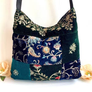 May include: A patchwork shoulder bag with a dark green strap. The bag is made of velvet in shades of dark green and navy blue, with embroidered floral designs in gold, red, and blue. The bag has a rectangular shape.